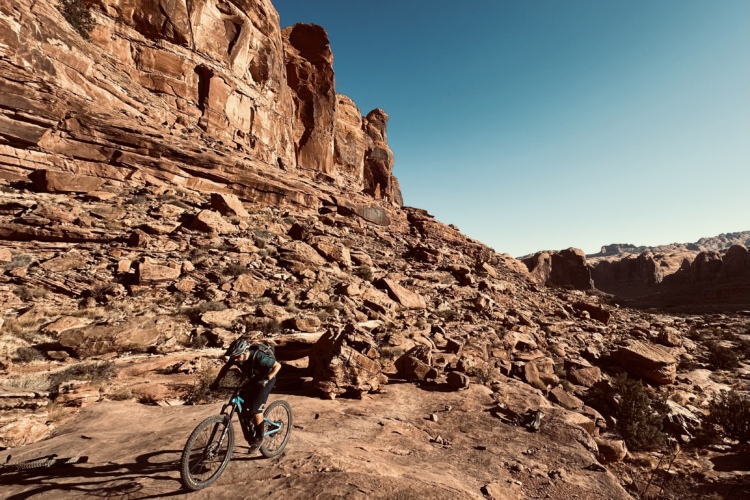 A mountain biker navigates a rocky landscape under a clear blue sky, surrounded by towering red rock formations. The terrain is rugged and uneven, showcasing natural textures and colors of the desert scenery.
