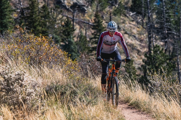 Lennard Zinn wearing a colorful jersey and helmet rides an orange mountain bike on a narrow, winding trail surrounded by tall grass and sparse shrubs, with trees and rocky terrain in the background.