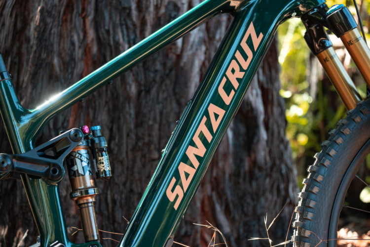 A close-up image of a Santa Cruz mountain bike frame, featuring a shiny green finish with prominent branding and details of the rear shock system. The background includes a blurred tree trunk and greenery, enhancing the outdoor setting.