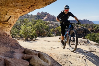 A mountain biker navigating a rocky trail surrounded by trees and rugged terrain, with a clear blue sky in the background. The scene captures the adventurous spirit of outdoor cycling. Eagle Eye mountain bike trail.