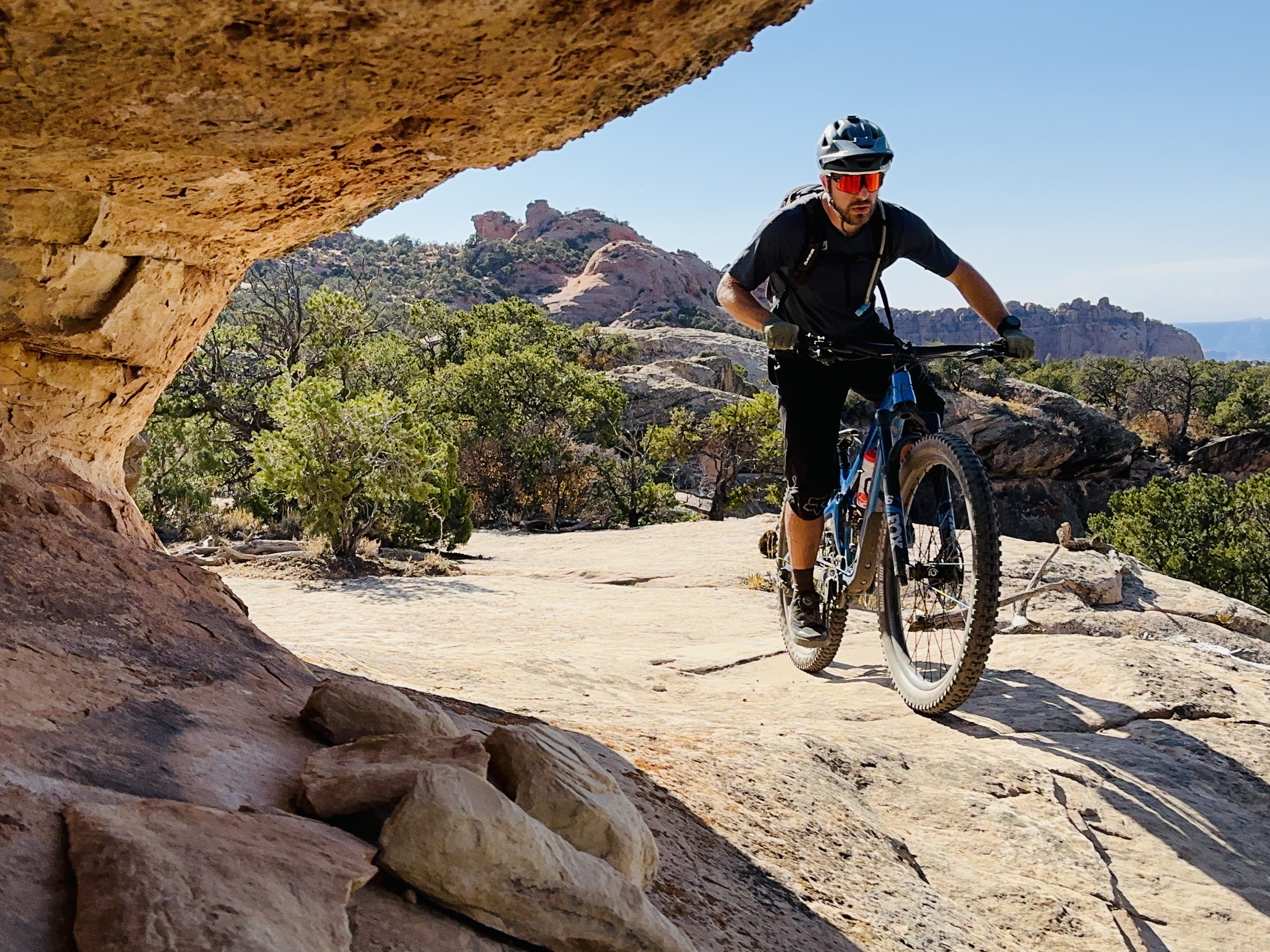 A mountain biker navigating a rocky trail surrounded by trees and rugged terrain, with a clear blue sky in the background. The scene captures the adventurous spirit of outdoor cycling. Eagle Eye mountain bike trail.