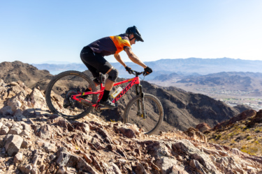A mountain biker riding a bright pink bike on rocky terrain at a high elevation, with a mountainous landscape and clear blue sky in the background. The rider is wearing a black helmet and a colorful cycling jersey. Bootleg Canyon mountain bike trail.