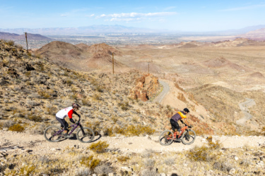 Two mountain bikers navigate a winding dirt trail through a rocky desert landscape, with rolling hills and sparse vegetation in the background. The sky is clear, and distant mountains are visible under the bright sun. Bootleg Canyon mountain bike trail.