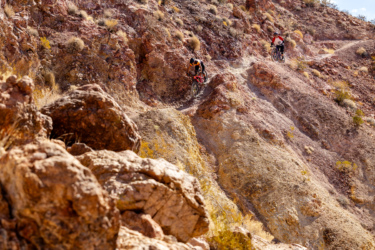 Two mountain bikers navigate a rocky, uneven trail through a rugged landscape. The terrain features large boulders and dry, earthy tones, with scattered vegetation in the background. One biker is dressed in an orange and black outfit, while the other is in a red and white jersey. Bootleg Canyon mountain bike trail.