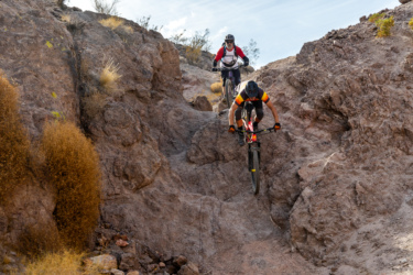 Two mountain bikers navigate a rocky terrain in a desert landscape. One cyclist is in the foreground, airborne while descending a slope, and the other is positioned higher up on the trail, preparing to follow. The rocky surface features sparse vegetation and dry shrubs, with a clear blue sky visible overhead. Bootleg Canyon mountain bike trail.