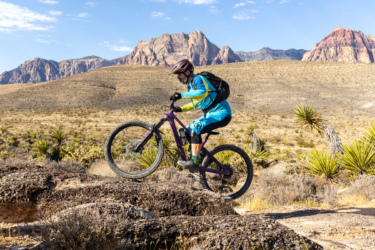 A mountain biker wearing a blue and yellow outfit and a helmet rides over rocky terrain, with a desert landscape and mountains in the background. The biker is in mid-action, lifting the front wheel off the ground. Cowboy Trails mountain bike trail.