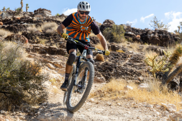 A mountain biker in a vibrant, striped jersey navigates a rocky trail, wearing a helmet and sunglasses. The sun shines down on a rugged terrain with dry vegetation and unique rock formations in the background. Cowboy Trails mountain bike trail.