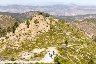 Four mountain bikers are riding on a trail through a lush green landscape, surrounded by rocky formations and distant mountains. The scene showcases a clear sky and expansive views of the hilly terrain, emphasizing the beauty of the outdoors. Ella Mountain Trail mountain bike trail.