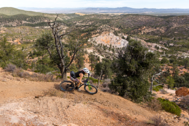 A mountain biker navigating a winding dirt trail on a rocky hillside, surrounded by trees and expansive views of a mountainous landscape in the background. The biker is wearing a helmet and cycling gear, highlighting the active outdoor environment. Ella Mountain Trail mountain bike trail.