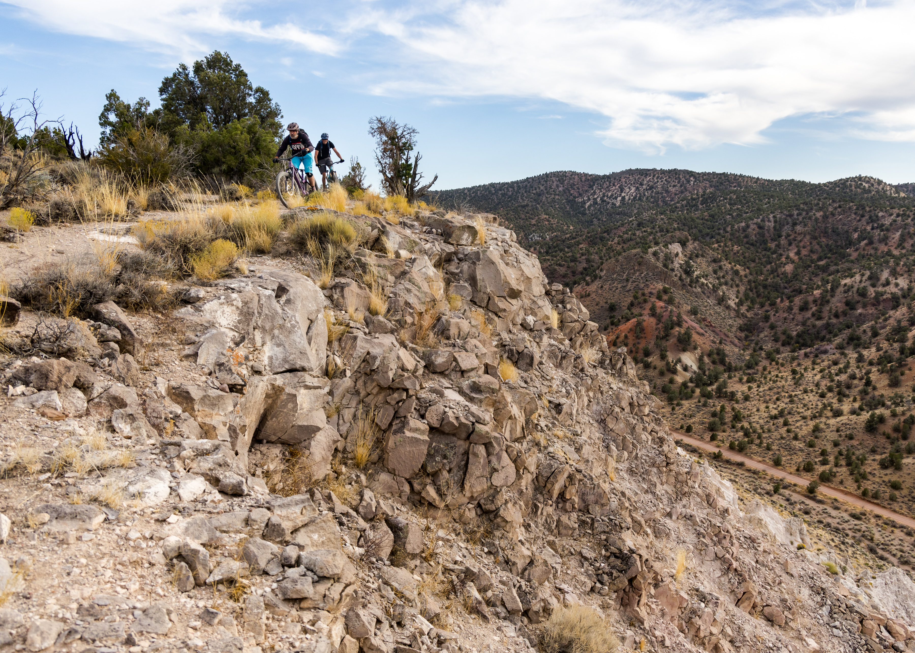 Two mountain bikers navigate a rocky trail along a cliff edge, surrounded by sparse vegetation and rolling hills under a partly cloudy sky. The landscape features rugged terrain with a distant dirt road winding through the valley below. Rocky Top mountain bike trail.