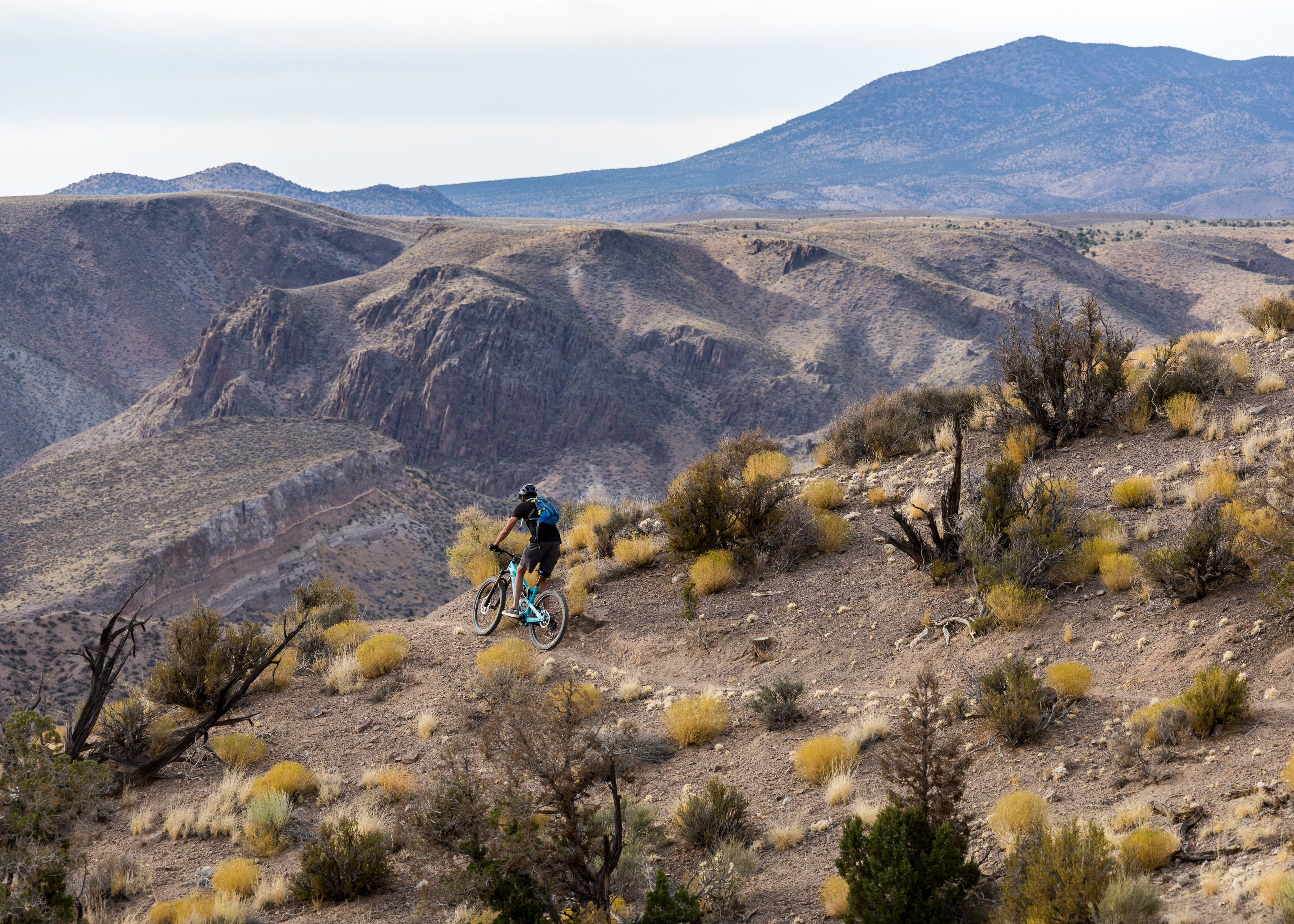 A mountain biker descends a rocky trail through arid terrain, with rolling hills and distant mountains in the background. The landscape features sparse vegetation, including small bushes and golden grasses, under a cloudy sky. Boar