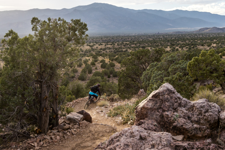 A mountain biker navigates a winding dirt trail surrounded by rocky terrain and greenery, with expansive hills and mountains in the background under a cloudy sky.