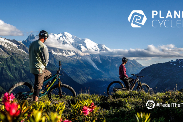 A scenic mountain landscape featuring two cyclists resting beside their bikes, with snow-capped peaks in the background and vibrant flowers in the foreground. The sky is clear with a few clouds, highlighting the natural beauty of the outdoor setting. The image includes the logo for "Planet Cyclery" in the top right corner and a hashtag "#PedalThePlanet" at the bottom.