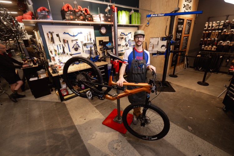 A bike mechanic in an apron stands in a workshop, holding a mountain bike on a repair stand. The background features a pegboard with various bike tools, parts on shelves, and a selection of bike tires, creating a bustling repair environment.