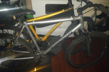 Diamondback Sorrento: A close-up image of a silver Diamondback mountain bike parked indoors, with a yellow bike partially visible in the background. The scene reveals a wooden floor and an appliance in the background, suggesting a home environment.