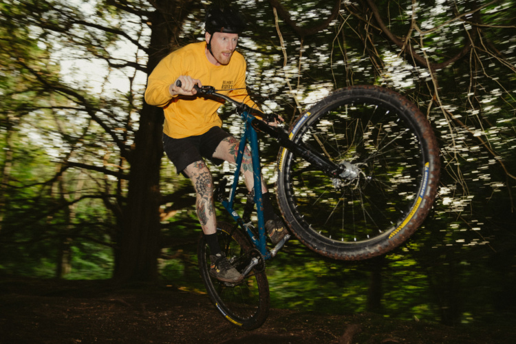 A mountain biker performing a jump on a trail surrounded by trees, wearing a yellow long-sleeve shirt and black shorts, with visible tattoos on his legs. The image captures the motion and energy of biking in a natural setting, with a blurred background emphasizing the speed of the rider.