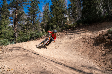 A person riding a mountain bike on a dirt trail through a forest, leaning into a turn. They are wearing a helmet and protective gear, with trees and blue sky in the background. Baker's Park mountain bike trail.