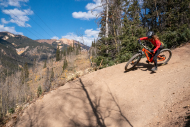 A cyclist riding down a dirt trail on a mountain bike, angled sharply on a hillside with trees and mountains in the background under a blue sky with fluffy clouds. Baker's Park mountain bike trail.