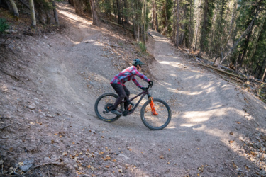 A mountain biker navigates a dirt trail through a forested area, leaning into a turn on a rocky, downhill path. The rider wears a helmet and a colorful plaid shirt, showcasing an active outdoor lifestyle surrounded by trees and natural scenery. Baker's Park mountain bike trail.