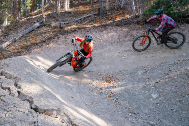 Two mountain bikers navigate a dirt trail in a forested area. One rider, wearing a red jersey and a helmet, leans into a turn on an orange bike. The second rider, dressed in a plaid shirt, follows closely behind on a black bike. The scene captures the excitement of mountain biking among trees with autumn foliage. Baker's Park mountain bike trail.