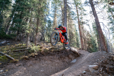 A mountain biker leaps off a dirt ramp in a forested area, surrounded by tall trees. The cyclist is wearing a helmet and a red and black jersey, demonstrating an aerial maneuver on a bright orange mountain bike, with dust kicking up from the trail below. Baker's Park mountain bike trail.