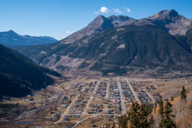 Aerial view of a small town nestled in a valley, surrounded by mountains under a clear blue sky. The town features colorful houses and streets, with a winding river visible nearby, and forests covering the slopes of the mountains that encircle the area. Baker's Park mountain bike trail.