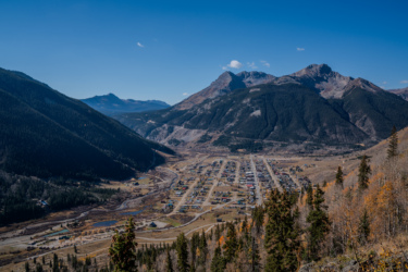 A panoramic view of a valley with a small town surrounded by mountains, featuring clear blue skies and scattered clouds. The town is nestled in the landscape, with winding roads and patches of greenery, while the backdrop showcases rugged mountains. Baker's Park mountain bike trail.