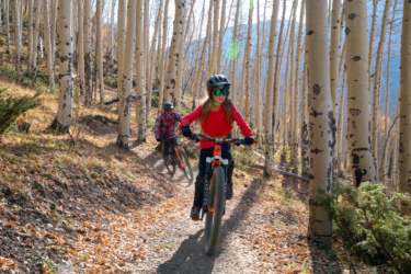Two mountain bikers ride along a narrow trail in a forest of tall aspen trees. One cyclist, a child wearing a red shirt and green sunglasses, leads the way, while an adult in a plaid shirt follows closely behind. The ground is covered in leaves, and sunlight filters through the trees, creating a vibrant, scenic atmosphere. Baker's Park mountain bike trail.
