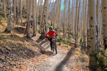 A child riding a mountain bike down a dirt trail surrounded by tall aspen trees. The scene captures autumn foliage with fallen leaves on the ground and sunlight filtering through the trees. Another cyclist can be seen in the background. Baker's Park mountain bike trail.
