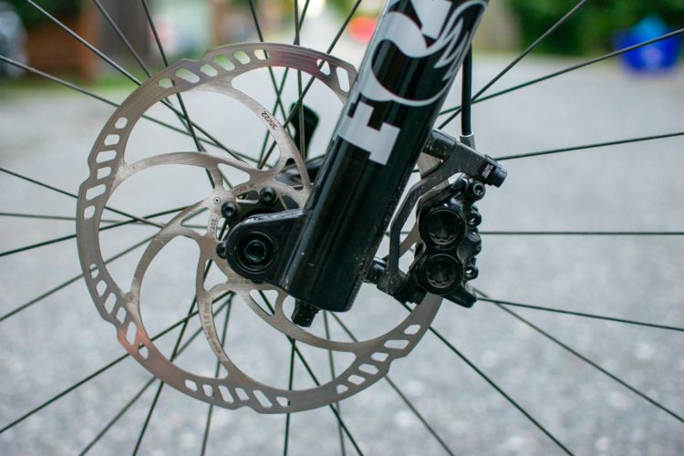 Close-up view of a bicycle's disc brake system, showcasing the rotor, brake caliper, and part of the fork on a gravel road surface.