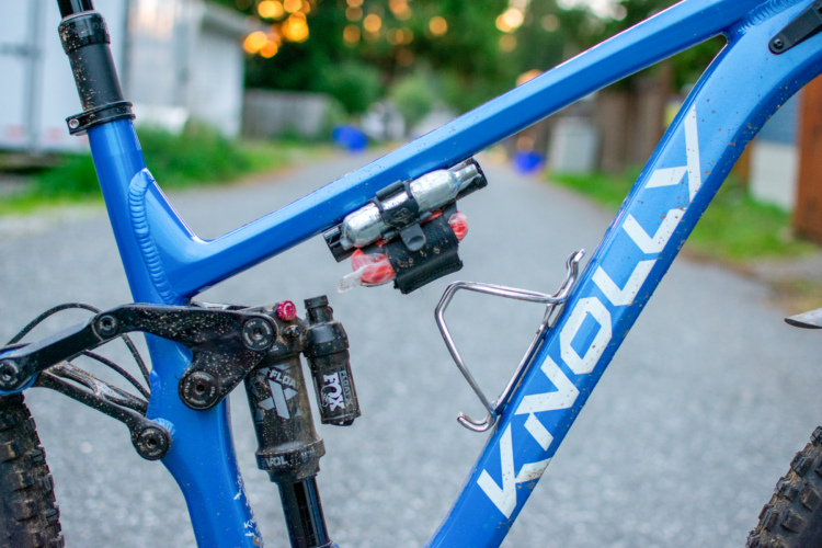 Close-up view of a blue Knolly mountain bike frame showcasing the suspension system, a water bottle cage, and a mounted CO2 cartridge holder. The bike appears to be used, with dirt and dust on the components, set against a gravel path.