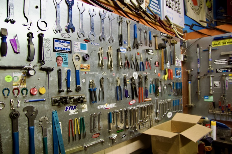 A detailed view of a workshop wall displaying an organized array of various hand tools, including wrenches, pliers, and screwdrivers, mounted on a metallic pegboard. A cardboard box is situated on the floor, and various stickers and business cards are visible among the tools.