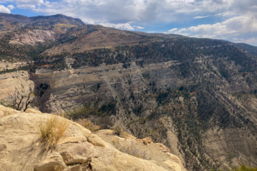 A panoramic view of rugged mountains featuring layered rock formations, with patches of greenery and sparse vegetation. The sky is partially cloudy, showcasing shades of blue and gray, while the foreground includes rocky terrain and dry grass. Palisade Plunge mountain bike trail.