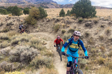Three mountain bikers navigate a winding dirt trail through a grassy landscape with sparse vegetation. The backdrop features rolling hills and mountains under a partly cloudy sky. Two riders are visible in the foreground, one dressed in bright blue and yellow gear, while the other wears a red jacket. A third rider can be seen in the distance, making their way around a curve in the trail. Palisade Plunge mountain bike trail.