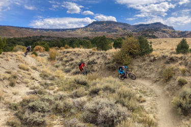 Three mountain bikers navigate a winding dirt trail surrounded by autumn foliage and rugged terrain, with mountains visible in the background under a partly cloudy sky. Palisade Plunge mountain bike trail.