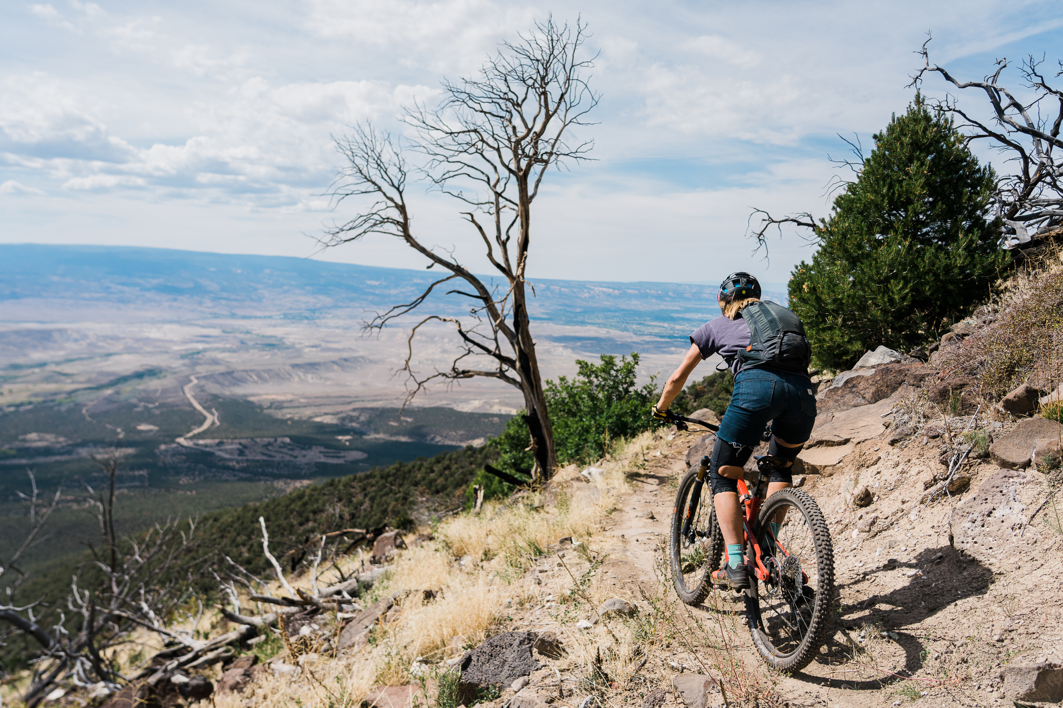 A mountain biker rides along a rocky trail with a scenic view of a valley below. In the background, a mix of dried and green trees lines the path, under a partly cloudy sky. The biker is equipped with a helmet and a backpack, navigating the rugged terrain. Palisade Plunge mountain bike trail.