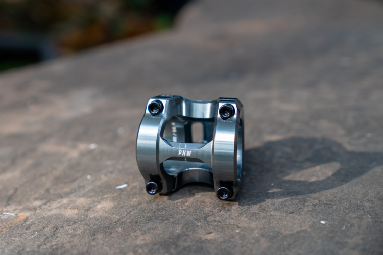 Close-up of a polished aluminum PNW Loam bicycle stem component, featuring six dark screws and a distinctive design with the letters "PNW" engraved on the side. The stem rests on a textured stone surface, with a blurred natural background.