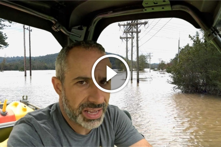 A person is sitting in a vehicle, speaking directly to the camera. The background shows a flooded area with standing water around power lines and trees. Some fuel containers are visible in the vehicle.