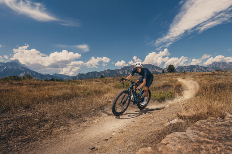 A cyclist riding a Mondraker Arid gravel bike in a blue outfit and white helmet navigates a dirt trail through a grassy area, with mountains and a blue sky filled with clouds in the background.