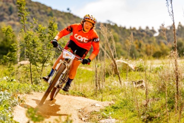 A mountain biker in an orange jersey and helmet navigates a winding dirt trail surrounded by greenery, showcasing determination and skill as they ride through a vibrant outdoor setting. Stromlo Forest Park mountain bike trail.