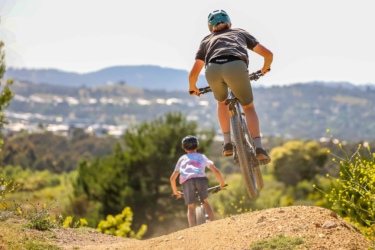 Two children riding bicycles on a dirt track in a green landscape. One child is jumping off a bump in the track, while the other trails behind, enjoying the ride. The background features trees and distant hills under a clear sky. Stromlo Forest Park mountain bike trail.