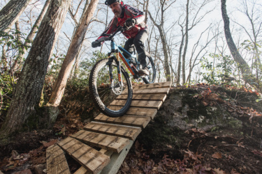 A mountain biker skillfully rides down a wooden ramp on a trail surrounded by trees and autumn foliage, capturing a moment of action and adventure in a forested setting. Satan's Ridge mountain bike trail.