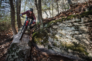 A mountain biker navigating a rocky trail surrounded by trees, balancing on a large stone while wearing a helmet and a flannel shirt. Leaves are scattered on the ground, indicating an autumn setting. Satan's Ridge mountain bike trail.