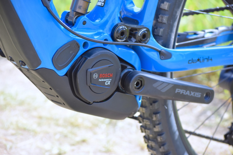 Close-up view of the bottom bracket area of a blue electric mountain bike, showcasing the Bosch Performance Line CX motor and Praxis crankset, with background elements blurred to emphasize the bike's components.