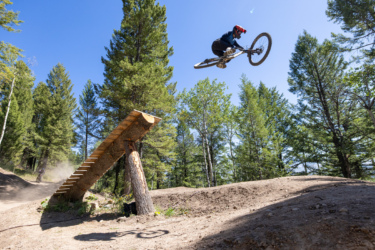 A mountain biker performing an aerial jump off a wooden ramp in a forested area, surrounded by tall trees and a clear blue sky. Dust is visible around the landing area, indicating an active trail. Deer Jumps mountain bike trail.