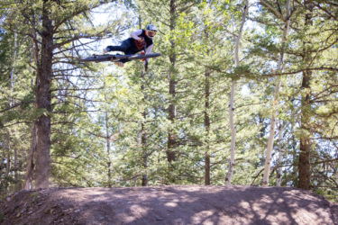A mountain biker performing a mid-air trick while jumping off a dirt ramp, surrounded by tall trees in a forest setting. The biker is wearing protective gear and a helmet, showcasing an action-packed moment in outdoor sports. Deer Jumps mountain bike trail.