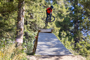 A mountain biker in a black helmet and red jacket performs a jump off a wooden ramp in a forested area. The bike is airborne, with trees and greenery in the background. Deer Jumps mountain bike trail.