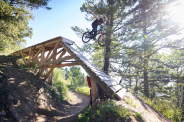 A mountain biker jumps off a wooden ramp situated in a wooded area, soaring through the air against a clear blue sky. The surrounding trees and greenery provide a natural backdrop, capturing the thrill of the ride. Deer Jumps mountain bike trail.
