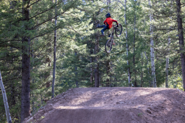 A person in a red shirt and helmet performs a mid-air trick on a mountain bike above a dirt jump, surrounded by dense green trees in a forested area. Deer Jumps mountain bike trail.