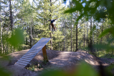 A cyclist performs a jump off a wooden ramp in a forested area, surrounded by tall trees. The rider is mid-air, showcasing an adventurous mountain biking moment. Deer Jumps mountain bike trail.